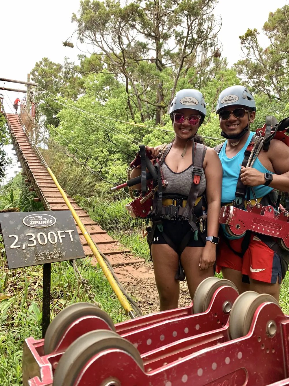Two guests in full zipline harnesses smile beside a Kapalua Ziplines trail sign marking a 2,300-foot line, with a suspension bridge visible in the forest behind them.