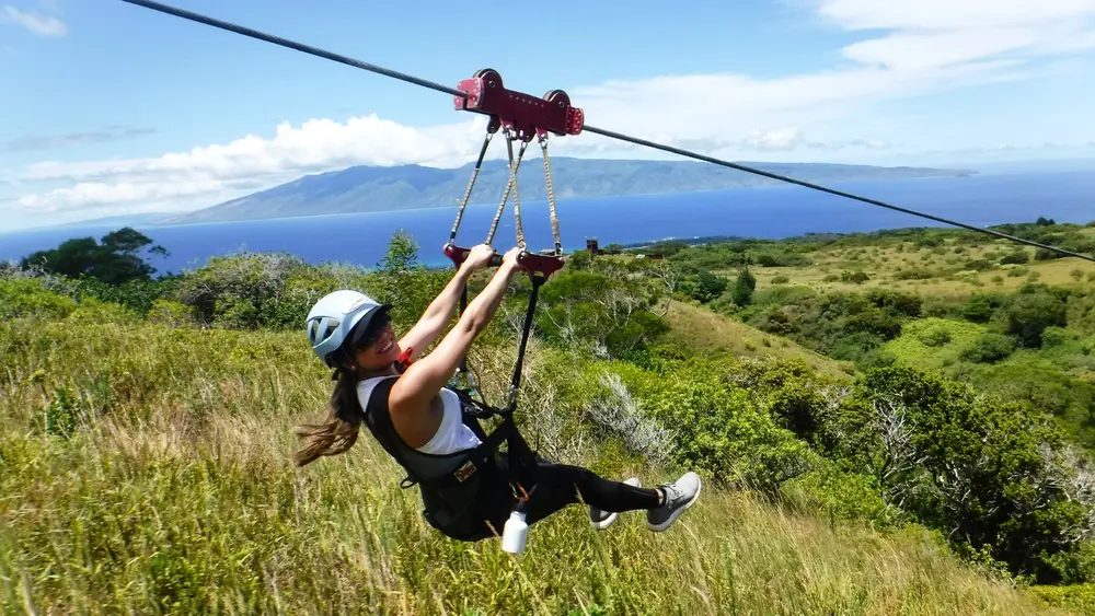 Guest smiling while ziplining over green hills, with the West Maui coastline and open ocean visible in the background.