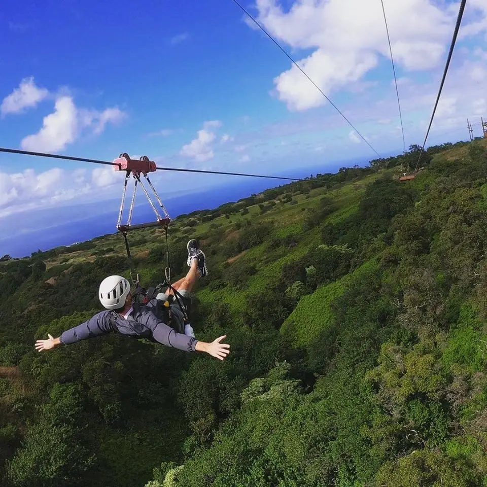 Guest launching from a zipline platform with arms spread wide, soaring above a forested Kapalua ridge with the ocean below.