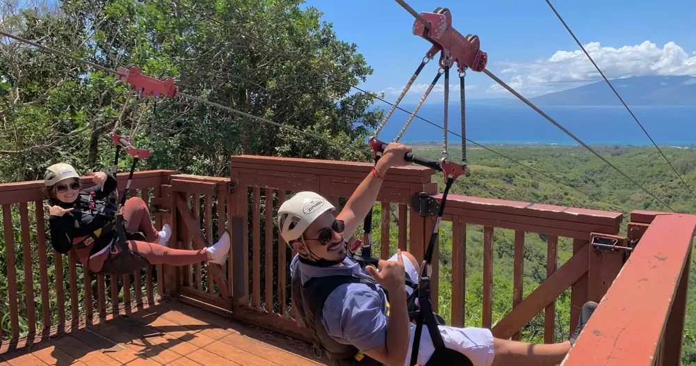 Two helmeted zipline riders prepare on a wooden platform high above a green Maui landscape, with the blue Pacific Ocean and neighboring island visible beyond.