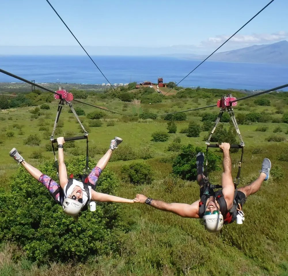 Two zipline riders hang upside down side by side over green Maui hillside, holding hands, with the Pacific Ocean and coastline in the background.