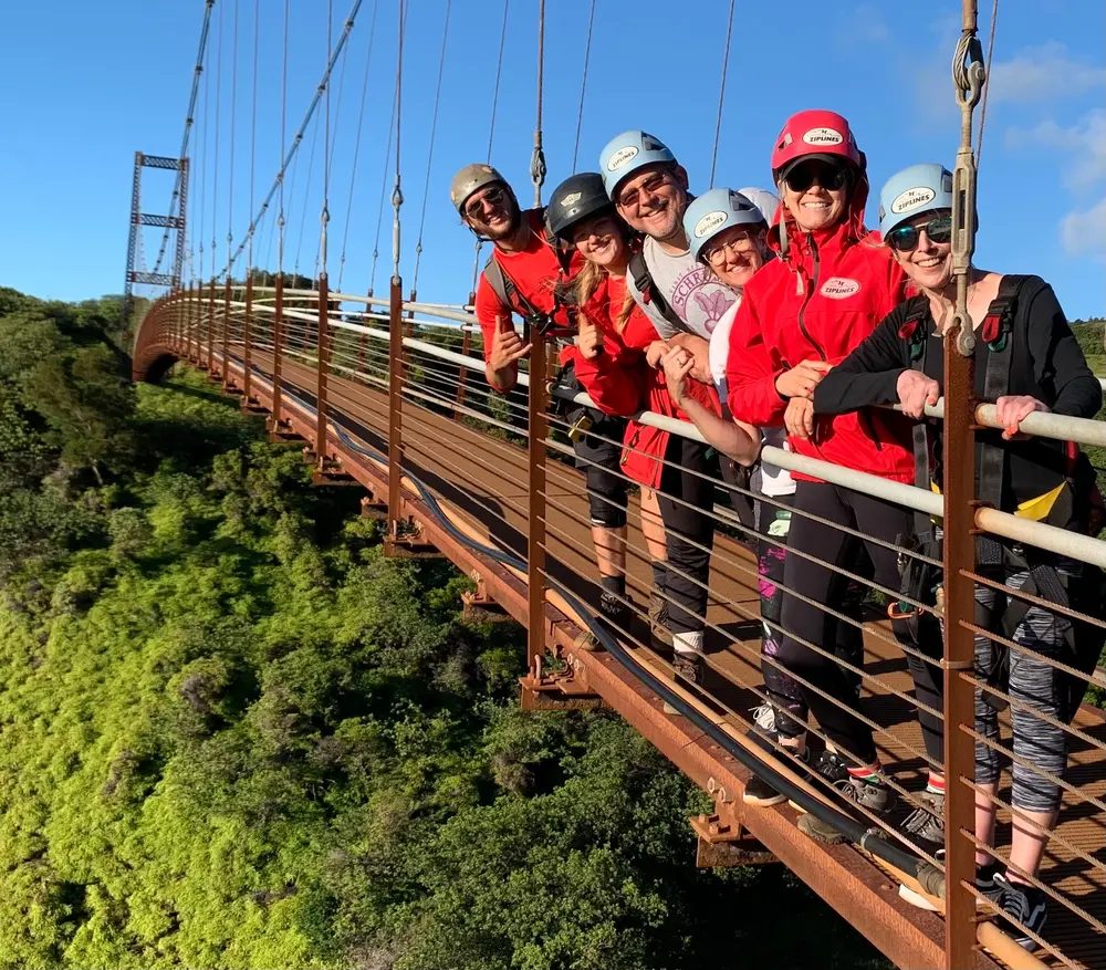Group of guests in helmets and harnesses smiling on the 360-foot suspension bridge, high above a lush green Maui ravine.