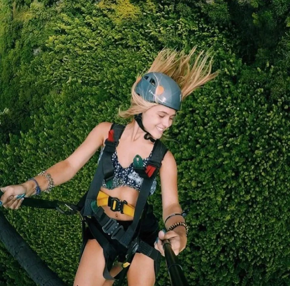 Close-up of a smiling zipline rider in a harness and helmet, hair blowing in the wind, with dense green foliage below.