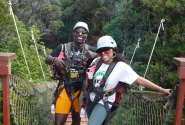 Two guests in helmets and harnesses crossing the suspension bridge together, smiling, surrounded by trees.