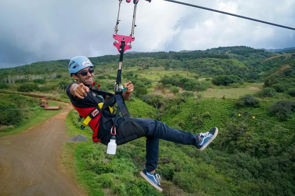 Zipline rider pointing at the camera mid-line, with a dirt access road and rolling green Maui hillside below.
